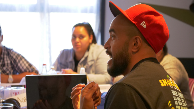 Māori man wearing red baseball cap seated at table with other colleagues