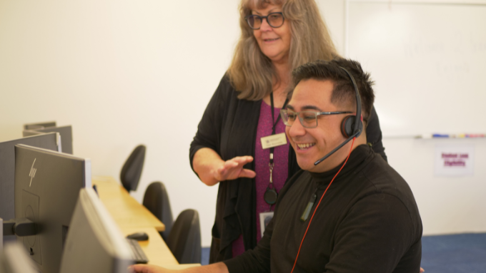 Man sitting at computer wearing headset, woman standing beside him. 