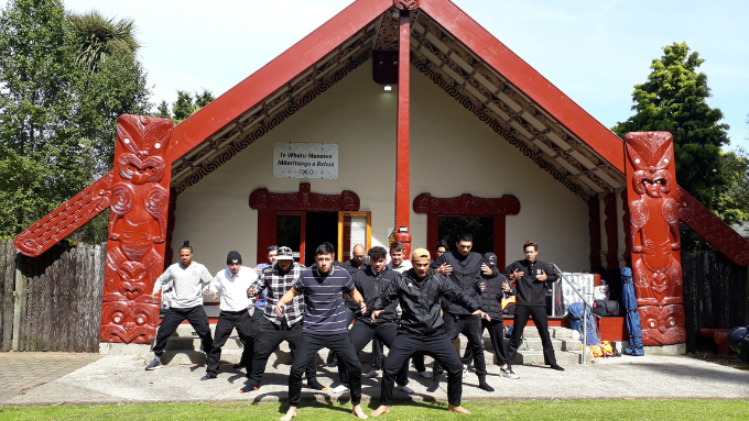 group of males preforming a haka infront of a marae behind them