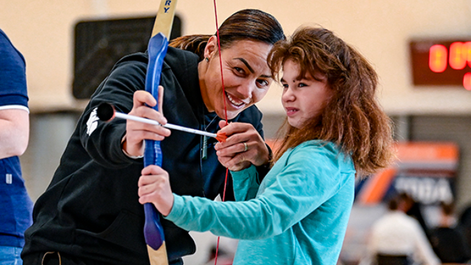 woman teaching a little girl how to shoot a bow and arrow indoors