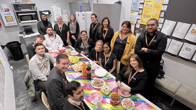 ASB staff in a lunch room with a table of food some seated some standing celebrating Pride week
