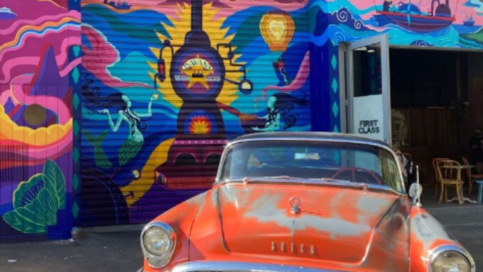 A beaten up old orange car against a colourful spray painted mural on a garage door