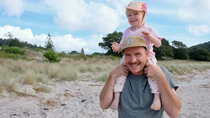 A man wearing a cap on a beach with a small child wearing pink on his shoulders both smiling