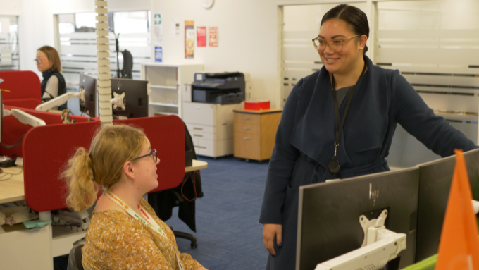 two woman talking to each other one seated at a desk, the other standing in an office two woman talking to each other one seated at a desk, the other standing in an office