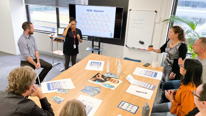 Southern Cross Travel Insurance staff in a meeting sitting on bar stools around a high table listening to a presenter with a large tv screen in the background