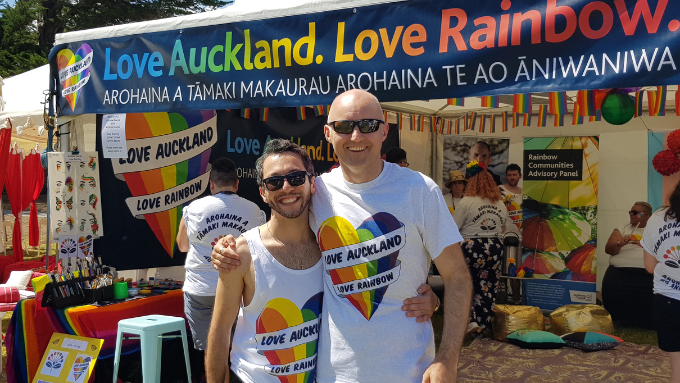 Two men wearing white t-shirts with the words Love Auckland and a rainbow heart standing in front of a Love Auckland. Love Rainbow market stand