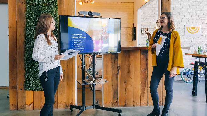 two women talking to each other standing in front of a large screen in a funky industrial office