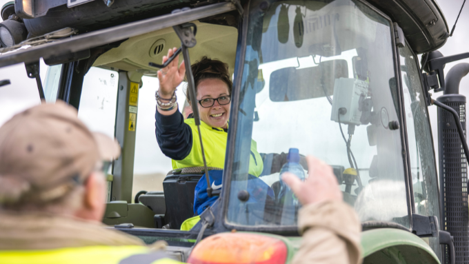 women inside the cab of a tractor waving out back towards co worker and camera smiling women inside the cab of a tractor waving out back towards co worker and camera smiling