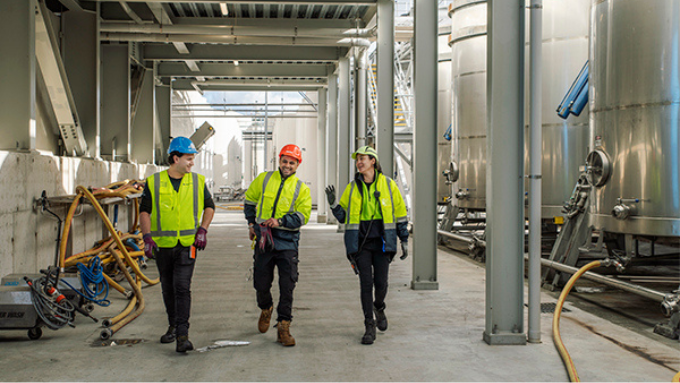 three workers talking and laughing wearing hi-vis vests and hard hats walking through a factory with large vats