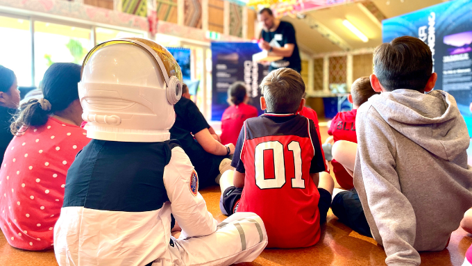 a man is talking to a group of children seated on the floor with their backs to the camera and one of them is wearing an astronaut costume 