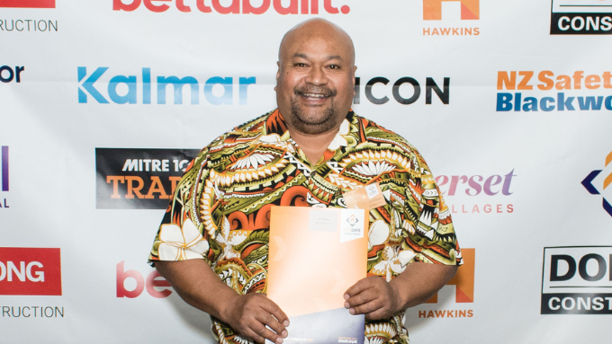 Site Safe New Zealand islander male infront of a brand board holding a booklet smiling looking at the camera