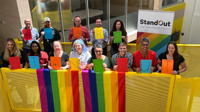 group photo of people holding up different coloured letters spelling we are standing out while encouraging pride
