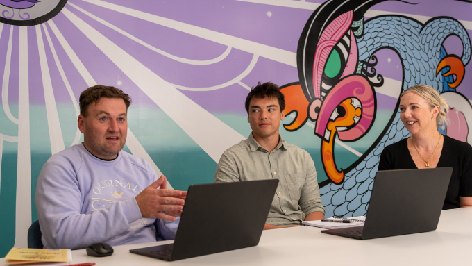 three people sitting at a table with laptops explaining something with a bright mural on the wall of a taniwha behind them