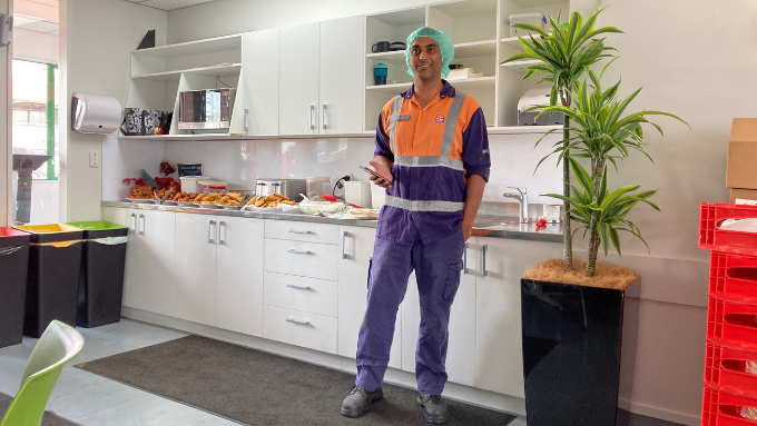 male worker standing in a kitchen smiling at the camera with food on the bench behind him