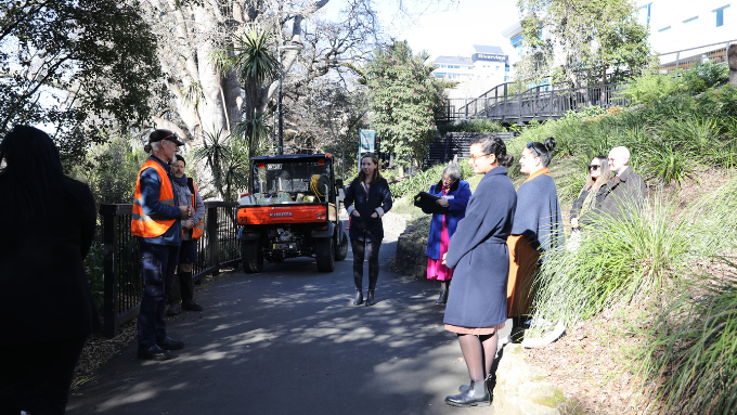 a group of people listening to two people in high vis vests on a walkway with native bush behind them