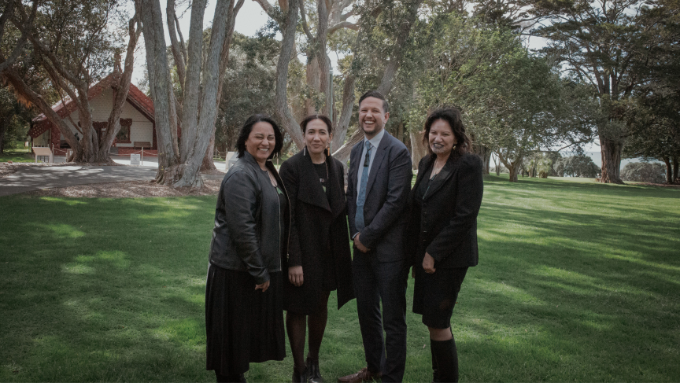 four people standing outside for a group photo with a marae and trees in the background