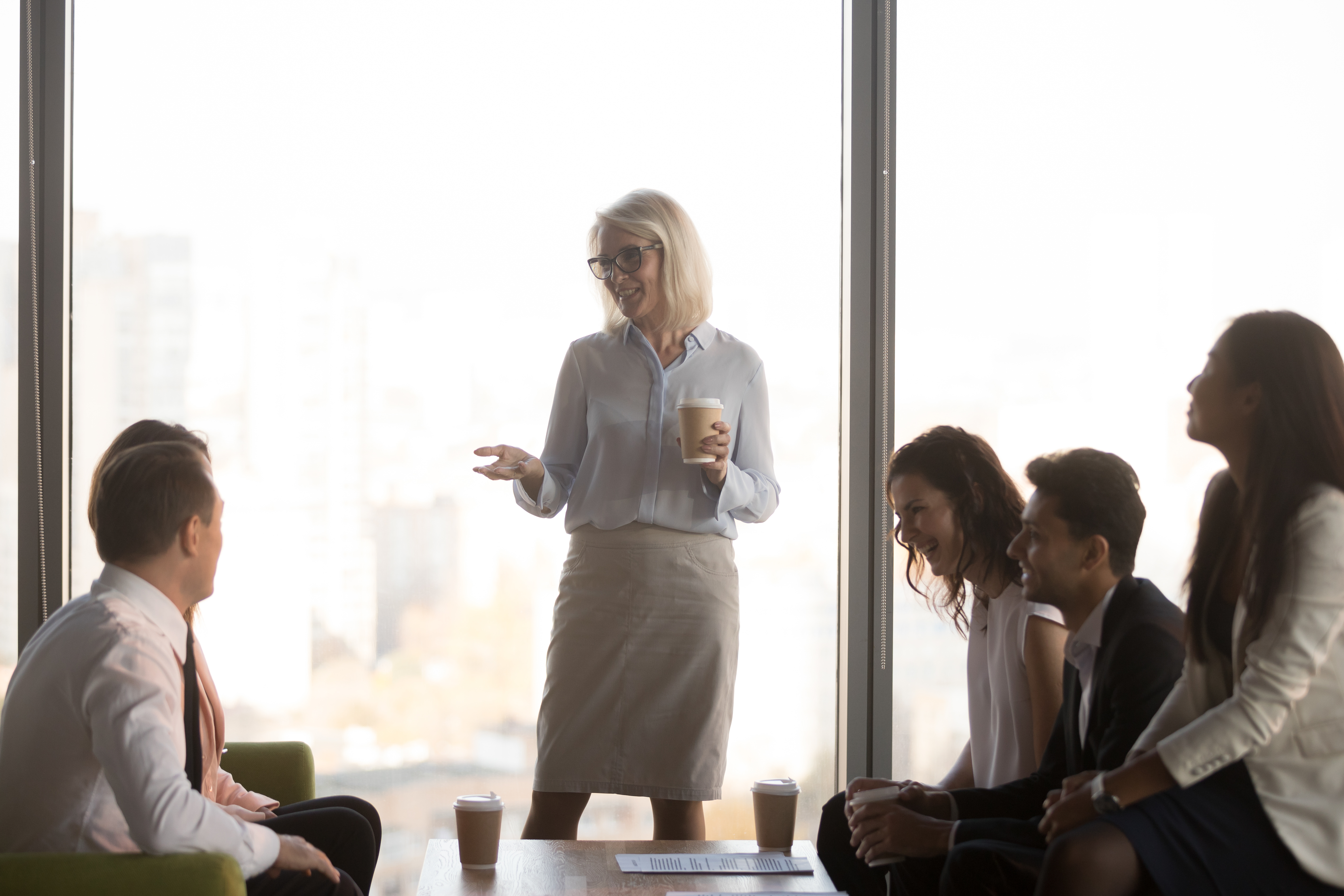 A woman standing in front of a window, with a coffee in her hand, presenting casually to a small group of people seated