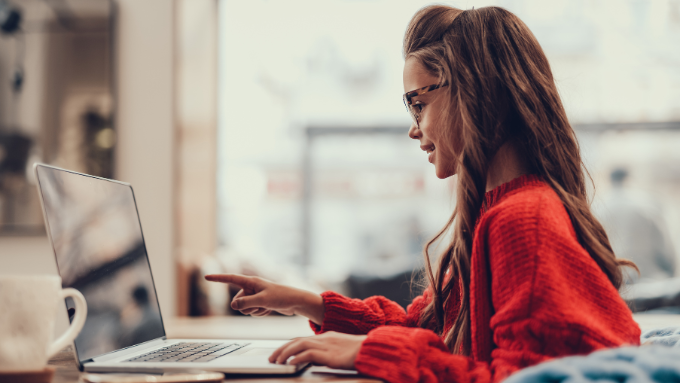 Engaged in her digital world, a young woman in a cozy red sweater type enthusiastically on her laptop. Sunlight filters through the café window, illuminating her determination and focus as she navigates her day.