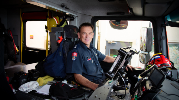 a fire fighter seated inside the cab of a fire truck facing the camera smiling 