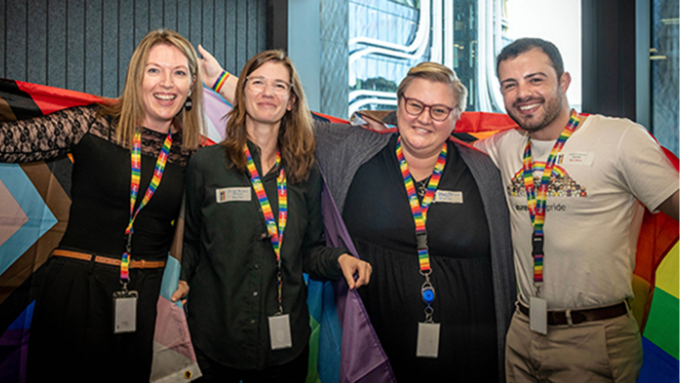 three women and one male smiling facing the camera with pride flags and colours and wearing rainbow coloured lanyards three women and one male smiling facing the camera with pride flags and colours and wearing rainbow coloured lanyards