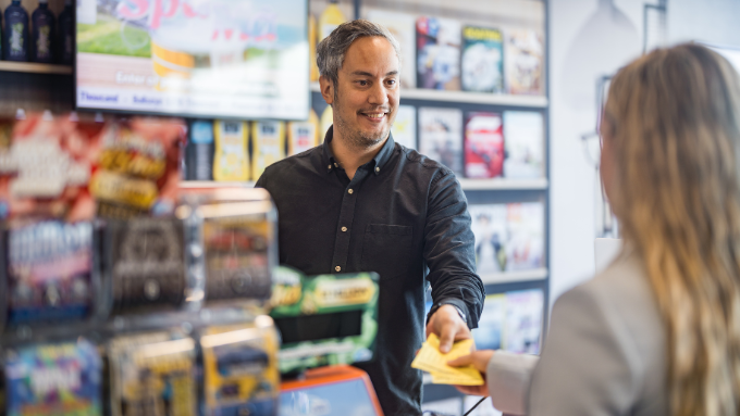 a woman buying a lotto ticket from a store with the owner smiling handing the ticket over 