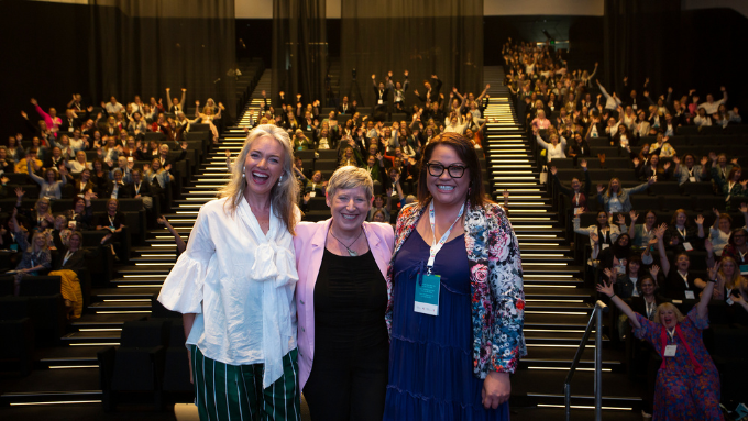 two tui raumata women and the MC of the event smiling facing the camera with a theatre crowd behind them seated two tui raumata women and the MC of the event smiling facing the camera with a theatre crowd behind them seated