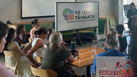 Group of people sitting in chairs in front of big screen with Te Uru Tāngata logo Group of people sitting in chairs in front of big screen with Te Uru Tāngata logo